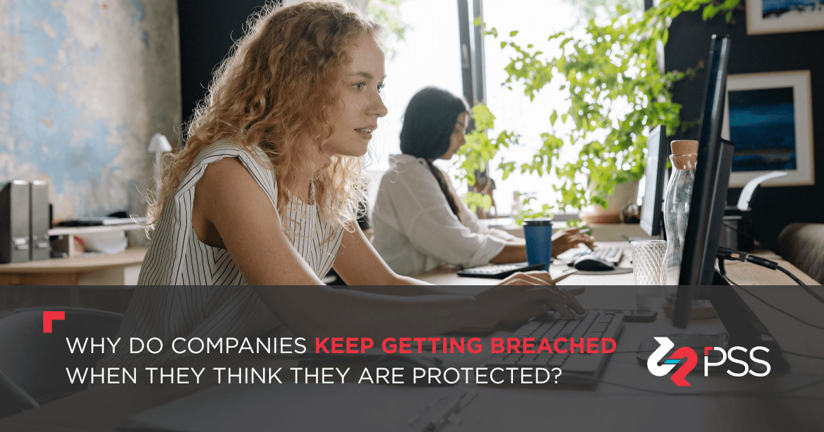 Two women working on computers in an office in front of a wide window. Text? Why Do Companies Keep Getting Breached When They Think They Are Protected